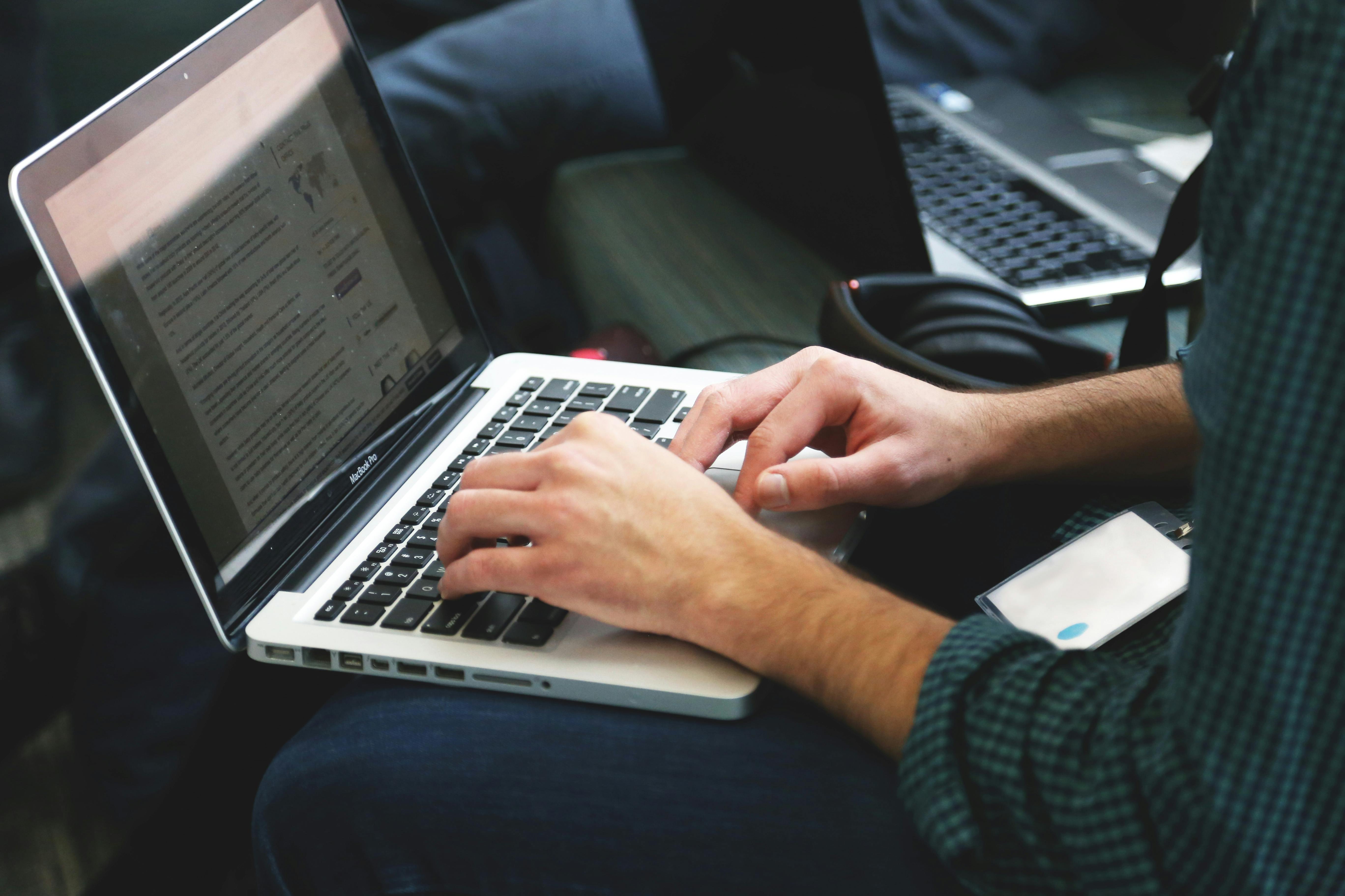 A man sits on a bench, typing on the laptop that sits on his lap.