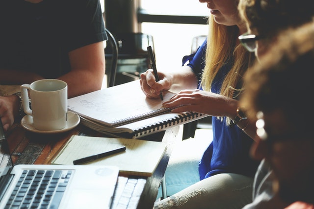 A group of people sit around a table with notebooks, coffee cup, and computer.